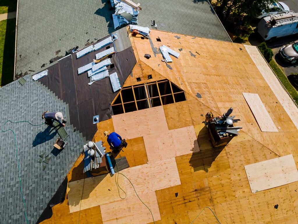 Roofing workers inspect exposed roof decking beside removed shingles on a residential house.